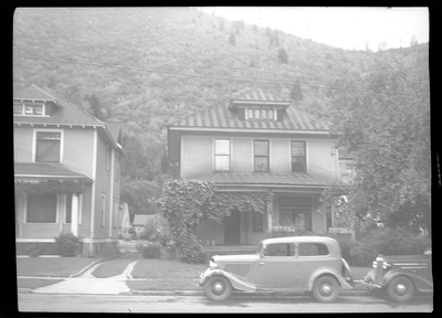 Photo of the Seelig house that has two cars parked on the side of the street in front of it. The house is two stories tall and has large trees in the front yard.