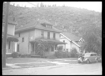 Photo of the Seelig house that has two cars parked on the side of the street in front of it. The house is two stories tall and has large trees in the front yard.