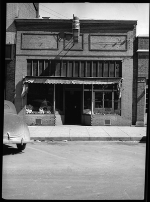 Photo of the storefront of Table Supply. There are two window displays on either side of the front door and one of the windows has several decals that read, "Table Supply," "Fruits," "Bakery," Meats," "Vegetables," "Groceries." There is a car parked in front of the building.