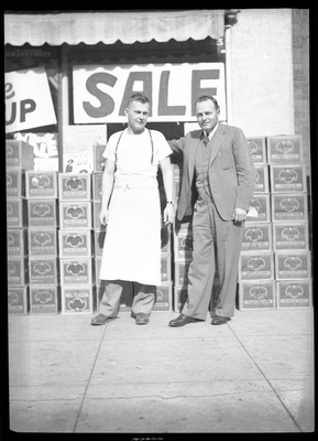 Two unidentified men stand in front of several stacks of boxes with a sign behind them that reads "sale." One man is wearing a suit and the other appears to be wearing an apron.