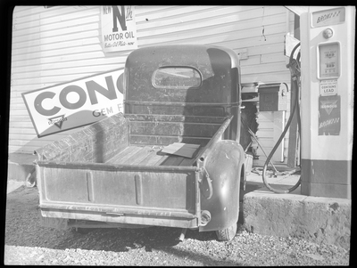 Photo of a truck that was crashed into the side of the General Food Store, damaging the wall and a sign on the exterior. There is a gasoline pump directly to the right of the truck, and a conoco sign fell on the hood.