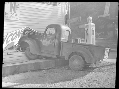 Photo of a truck that was crashed into the side of the General Food Store, damaging the wall and a sign on the exterior. There is a gasoline pump directly to the right of the truck, and a conoco sign fell on the hood.