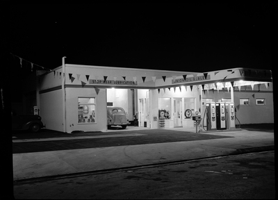 Photo of the Union Oil Dealer building at night. The store is still open with the lights on, and there is a car parked in the garage. Above the garage is a sign that reads, "Stop-Wear Lubrication" and outside there are three gas pumps.