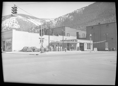 Photo of the Phillips 66 Gas Station in Wallace, Idaho. The gas station is on a street corner and has multiple gas pumps in front of it, as well as two garages attached to the building itself, with one of them open. There are three cars and a truck parked along the side of the building next to the gas station.