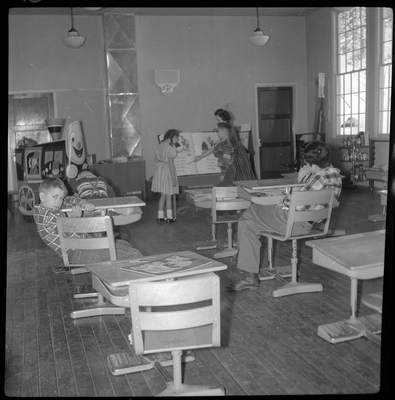 Photo of a classroom within the Opportunity School. Four kids and a teacher are visible; two kids and the teacher are in the back of the room and looking at an oversized book while the other two children sit in the school desks. One of the children is looking back at the camera.