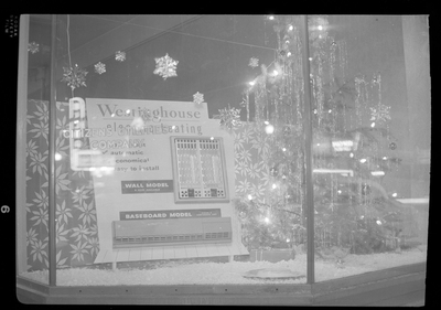 Photo of the Christmas themed window display inside the Citizens Utility building. There is a sparsely decorated Christmas tree and fake snow.
