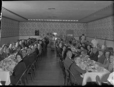 Photo of the dining room inside the Benevolent and Protective Order of Elks building. There are two lines of tables on either side of the room that have men and women seated at them, and a few waitresses are handing out food. Most people sitting at the tables are looking at the camera for the photo.