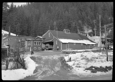 Photo of the exterior of the Wallace Meat Company building. It is connected to what appears to be a house or office building, which has a garage attached. There are several cars parked outside and there is snow on the ground. There are also two men outside.