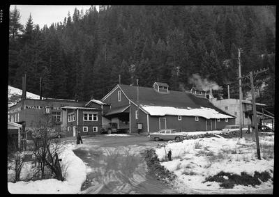 Photo of the exterior of the Wallace Meat Company building. It is connected to what appears to be a house or office building, which has a garage attached. There are several cars parked outside and there is snow on the ground. There are also two men outside.