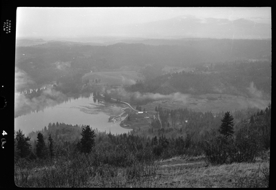 Scene looking over the land from the top of a hill or mountain. The photo is either looking down at Revelli Ranch, or taken from a view vantage point at Revelli Ranch. A lake and trees are visible, as is a handful of buildings on farmland.