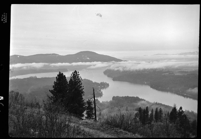 Scene looking over the land from the top of a hill or mountain. The photo is either looking down at Revelli Ranch, or taken from a view vantage point at Revelli Ranch. A lake and trees are visible, as is a handful of buildings on farmland.