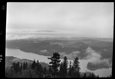Scene looking over the land from the top of a hill or mountain. The photo is either looking down at Revelli Ranch, or taken from a view vantage point at Revelli Ranch. A lake and trees are visible, as is a handful of buildings on farmland.