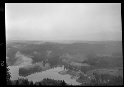 Scene looking over the land from the top of a hill or mountain. The photo is either looking down at Revelli Ranch, or taken from a view vantage point at Revelli Ranch. A lake and trees are visible, as is a handful of buildings on farmland.