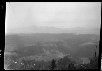 Scene looking over the land from the top of a hill or mountain. The photo is either looking down at Revelli Ranch, or taken from a view vantage point at Revelli Ranch. A lake and trees are visible, as is a handful of buildings on farmland.