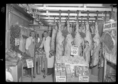 Photo of two unidentified men standing with a meat display inside City Meat Market. Sausage is listed at $0.59 per pound and bacon is listed at $0.55 per pound.
