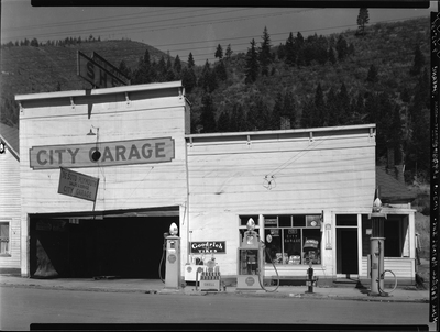 Photo of the exterior of the City Garage. There are several signs on the outside advertising products and services, as well as three gas pumps. The garage door is open.