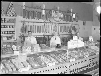 Three unidentified men stand behind the display counter at City Meat Market. There are sausages hanging from meat racks on the wall behind them, and raw meat in the display cases.