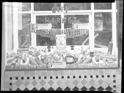 Photo of the window display at City Meat Market. There are various cured meats displayed in the window, and several signs that read, "Make it safe and sane; a frankly 4th; select these easy-to-serve July 4th meats from this display!"
