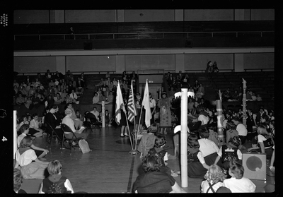 Group of unidentified people gathered in a room for the Blue Bird Fly-Up.Most people are facing away. Three flags are visible in the middle of the room.