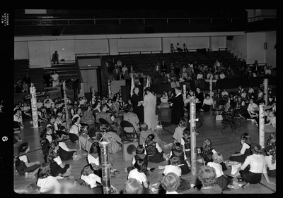Group of unidentified children gathered in a room for the Blue Bird Fly-Up. Children are sitting in a circle with three adults standing in the middle. There are decorated poles and flags visible.