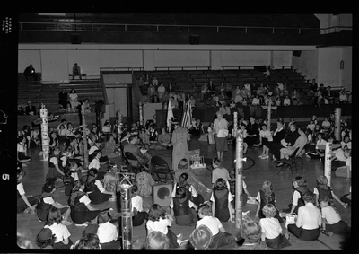 Group of unidentified children gathered in a room for the Blue Bird Fly-Up. Children are sitting in a circle and there are adults standing in the middle. There are decorated poles and flags visible.