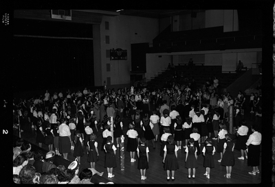 Group of unidentified people gathered in a room for the Blue Bird Fly-Up. Men, women, and mostly children are all standing in a circle. Decorated poles and flags are visible.