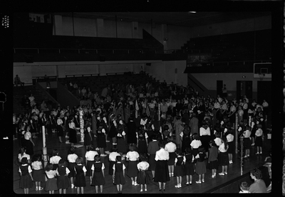 Group of unidentified people gathered in a room for the Blue Bird Fly-Up. Men, women, and mostly children are all standing in a circle. Decorated poles and flags are visible.