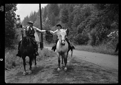 Two horse back riders, a man and a child, from Pony Express on a dirt road. They are both holding up the same bag between them.