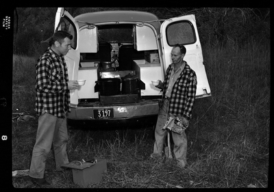 Two men from Shoshone County Search and Rescue stand at the back of the emergency vehicle while holding cups. The back of the car is open and it is parked in a grassy area. The license plate number is 3197.