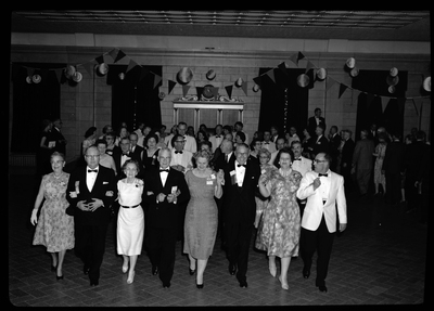 Lines of men and women walking into the Rotary International Club Convention, eight at a time. They are all holding hands or have their arms locked together. There are decorations strung up around the room and other people can be seen getting in line as well.
