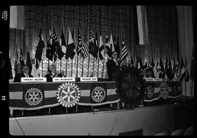 Rotary International Club Convention panel that includes Gordon Beaton, Guy M'Loughlin, Bill Featherstone, Theodore Hopf, Ira Branson, Jack Coventry, and Jack Barre. Some of the people listed are not behind their name placards, and there is an unidentified man speaking to the crowd. There are various flags along the back wall.