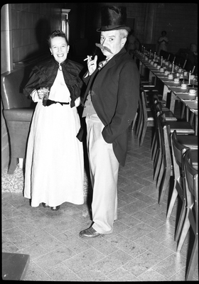 Photo of a man and woman at a Rotary Club party. They are both dressed nicely and the man is wearing a presumably fake mustache while smoking. The woman is holding a drink.