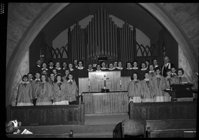 Staged photo of a church choir, possibly Methodist. Men, women, and children are lined up in rows and wearing matching robes. They are standing in front of what is likely an organ.