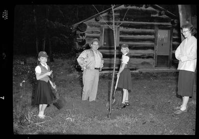 Photo of two young girls planting a tree with the help of a man and a woman. The two girls are wearing matching uniforms for Camp Fire Girls and they are both holding shovels. There is a log cabin behind them and they appear to be in a forest.