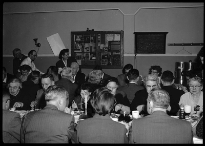 Photo of several groups of people seated at tables, eating together, for the 60th anniversary celebration of Fraternal Order of Eagles. Some people can be seen walking behind those seated. There is what appears to be a bulletin board on the wall behind everyone.