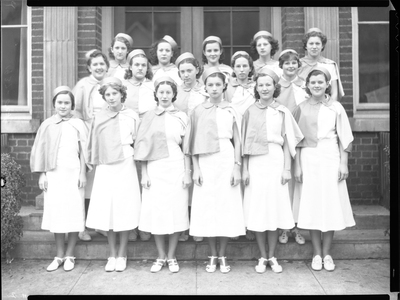 Photo of sixteen women standing together in three rows. They are wearing matching outfits and standing on the steps of a building for the photo. Previously described as "Theta Rho Girl's Drill Team."