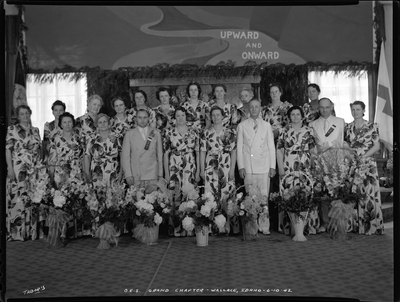 Group of women and men for the Order of Eastern Star Idaho Grand Chapter standing together. The women are all wearing matching dresses and the men are in suits, and there are bouquets of flowers on the ground in front of everyone. A flag can be seen in the corner, and the wall behind everyone reads "upward and onward."