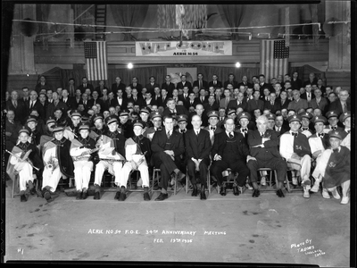Photo of a large group of men from the Fraternal Order of Eagles sitting with marching band members. Writing on the negative says: "Aerie No. 54 F.O.E. 34th Anniversary Meeting, Photo by Tabor's, Wallace, Idaho." There are American flags hanging in the background and a banner welcoming people to Aerie No.54.