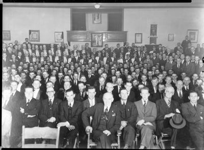 Photo of a large group of men from the Fraternal Order of Eagles sitting with marching band members. Writing on the negative says: "Aerie No. 54 F.O.E. 34th Anniversary Meeting, Photo by Tabor's, Wallace, Idaho."