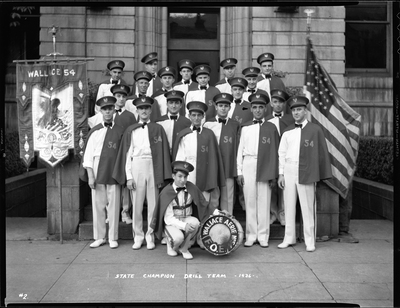 Group of men from the Fraternal Order of Eagles standing outside with a bass drum that has "Aerie No. 54 F.O.E." written on it. The negative has "State Champion Drill Team" written on it. The men are all wearing matching uniforms and there is a flag on either side of them.