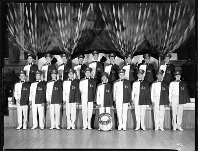 Group of men from the Fraternal Order of Eagles standing inside with a bass drum that has "Aerie No. 54 F.O.E." written on it. The men are all wearing matching uniforms with half of them standing on top of a stage behind the front row of men.
