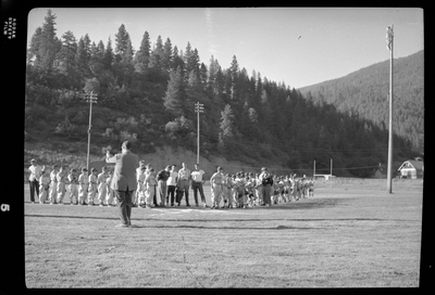 Two little league teams lined up on the field with their coaches. There is a man standing in front of them that appears to be speaking to them.