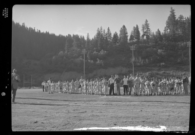 Two little league teams lined up on the field with their coaches. There is a man standing in front of them that appears to be speaking to them.