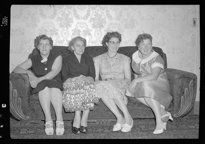 Four women sitting on a sofa together for photos. They are all wearing nice dresses.