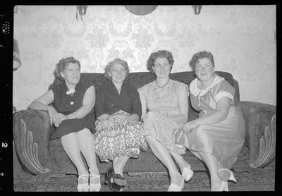 Four women sitting on a sofa together for photos. They are all wearing nice dresses.