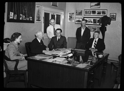 Five men and a woman are sitting a desk, looking at paperwork together. there are pictures and posters on the walls behind them. Previously described as "Ellis Hale in Coeur d'Alene Hardware Office."
