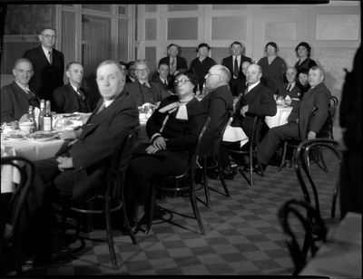 Group of finely dressed men and women (mostly men) sitting or standing around a table. Most of the people in the photo are looking at the camera.