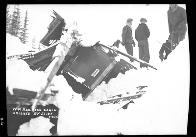Photo of a Northern Pacific Railroad coach after it was crushed by an avalanche. Some people can be seen standing next to the damaged train coach surveying the damage. Three people died.