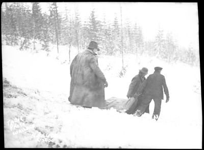 Three people are walking down a snow covered decline, two appear to be dragging something behind them. They are responding to an avalanche hitting a train on the Northern Pacific Railroad. Photos say that three people died.