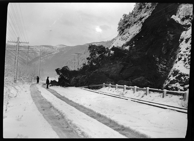 Two people stand at the base of a landslide that fell into the roadway on the Wallace-Kellogg Highway. There is snow on the ground, but it does not appear to be an avalanche. Previously described as "Rock slide, Rocky Point, 1 mile west of Wallace, Idaho."
